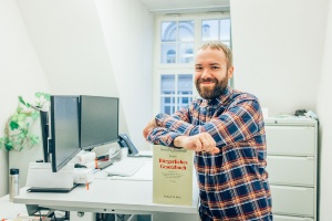 Portrait Duales Studium Bachelor of Laws © Philipp Marthaler Ein Auszubildender steht vor seinem Büroschreibtisch und lehnt sich mit den Armen auf ein Gesetzbuch