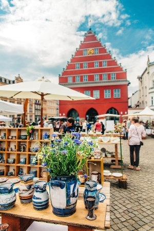 Markt in Greifswald © Wally Pruß Auf dem Historischen Marktplatz findet ein Sondermarkt statt. Im Vordergrund ist ein Frühlingsstrauß Blumen und ein Töpferstand zu sehen, im Hintergrund schlendern Gäste über den Markt.