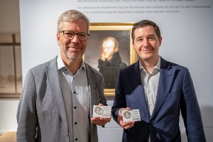 Dr Stefan Fassbinder und Lars Knevels von EuroMint zeigen die Jubiläumsmünze vor dem Porträt von Caspar David Friedrich im Pommerschen Landesmuseum